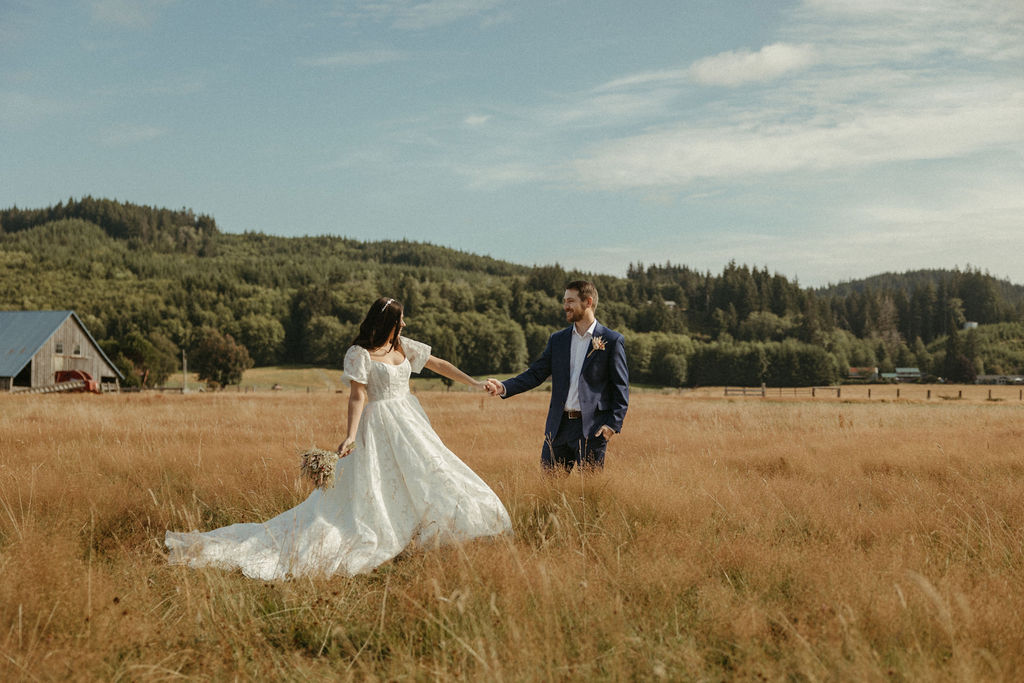 Bride and groom stand hand-in-hand in a golden pasture with towering pines and a barn in the background